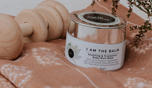 pink blanket and woode rattle in background of baby bum balm, white jar, silver lead, white label with botanical logo