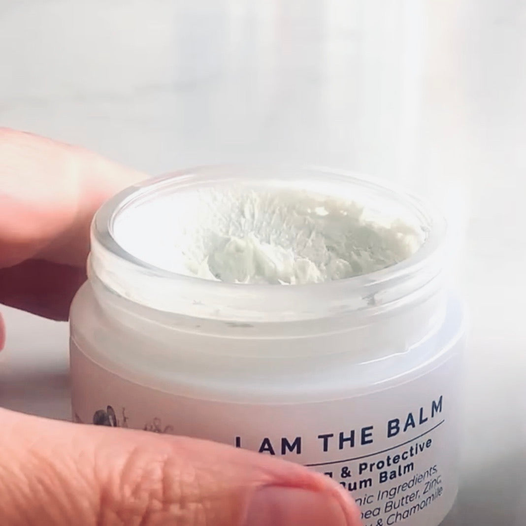 pink blanket and woode rattle in background of baby bum balm, white jar, silver lead, white label with botanical logo